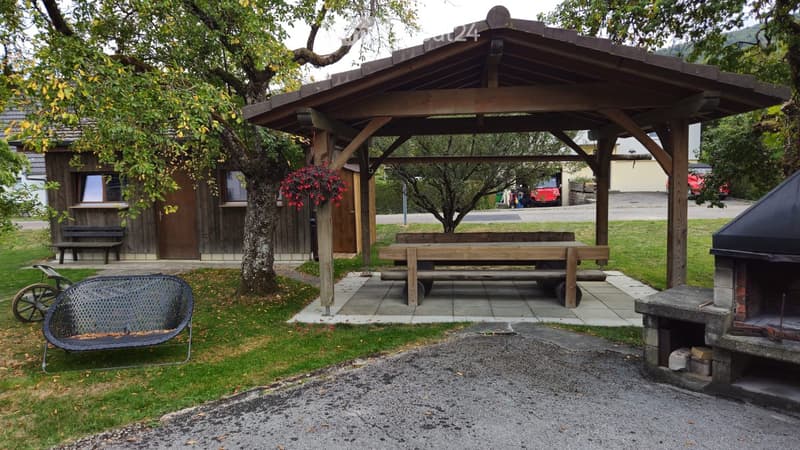 Outdoor seating area with a wooden table, benches, a barbecue, and a swing, surrounded by trees and a small wooden structure.