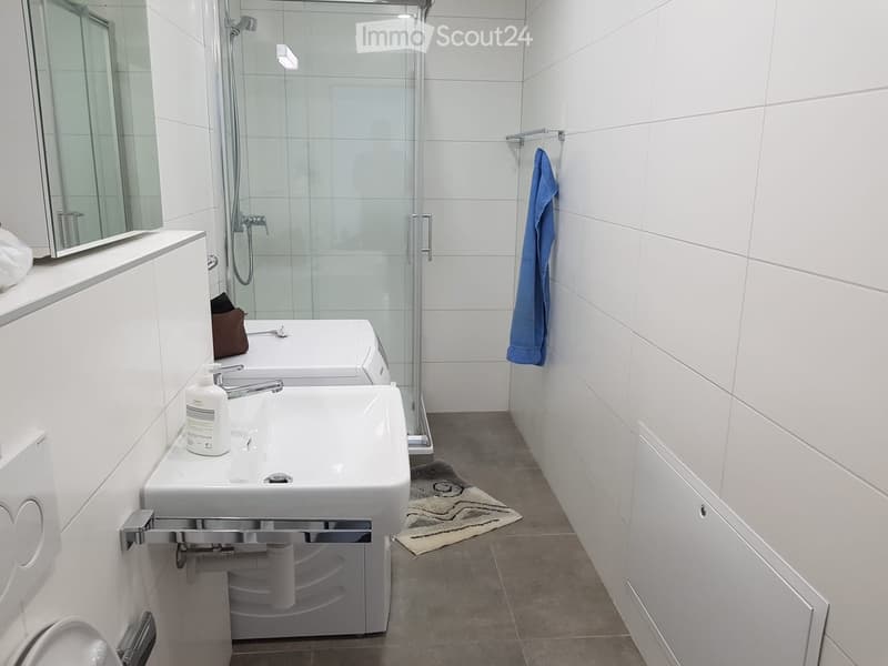 Modern bathroom with white tiles, a double sink, a glass-enclosed shower, a mirror, and a towel hanging on the wall.