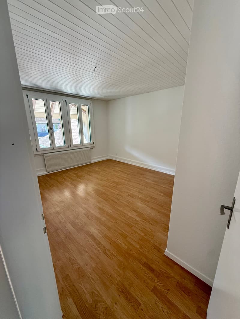 Empty room with wooden flooring, white walls, and large windows with white frames. A radiator is visible below the windows.