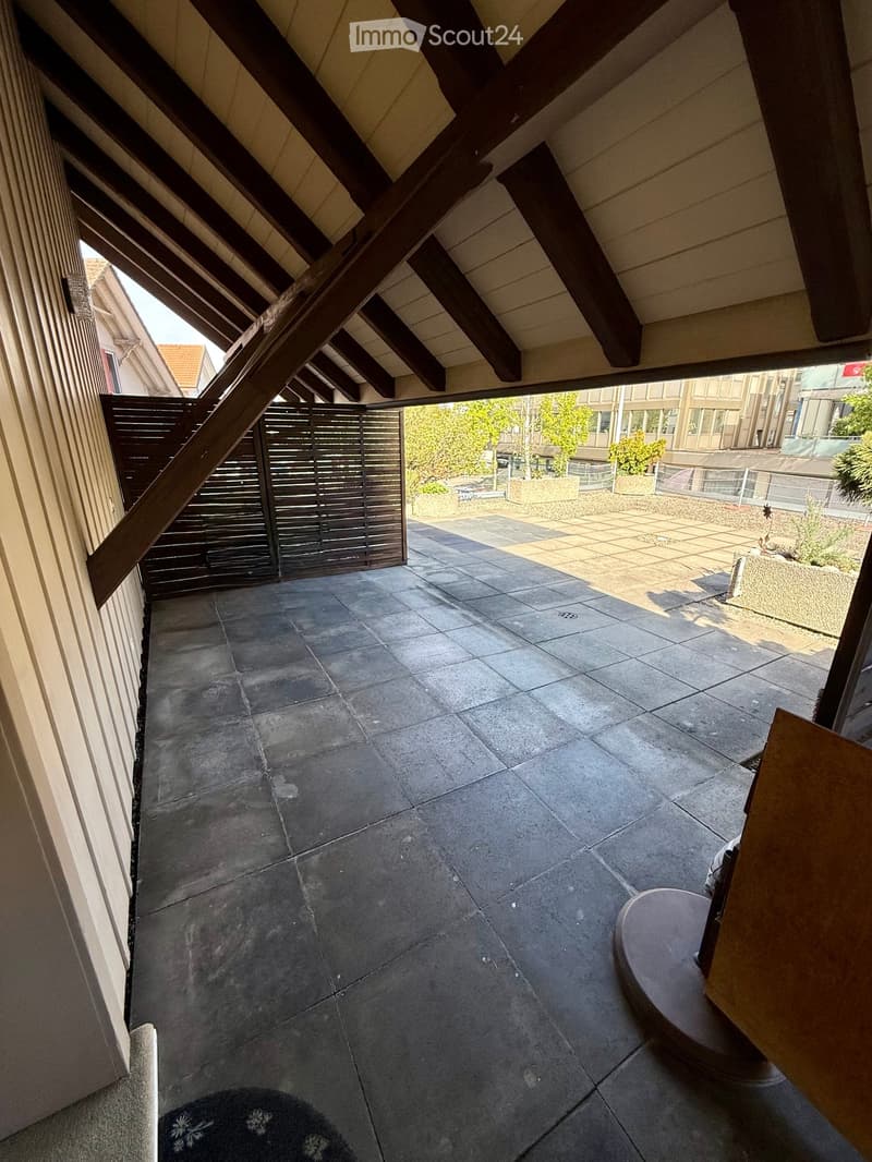 A covered balcony with dark tiled flooring, wooden beams, and a view of a garden area with plants and a paved patio.