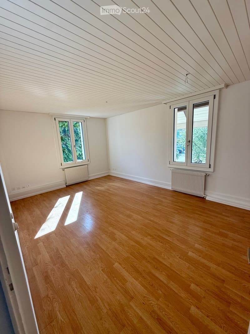 Empty room with wooden flooring, white walls, and large windows with white frames. Radiators are visible beneath the windows.