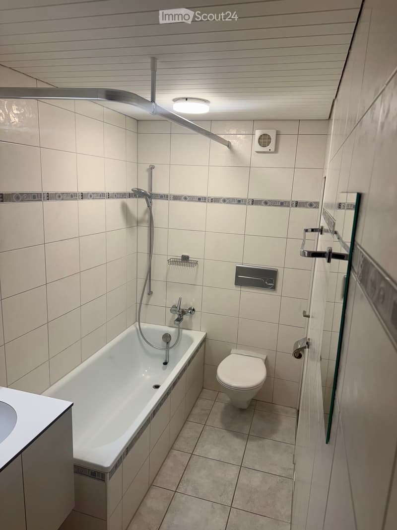 Modern bathroom with a bathtub, toilet, showerhead, and towel rack. The walls are tiled with a decorative border, and the floor is tiled with light-colored tiles.
