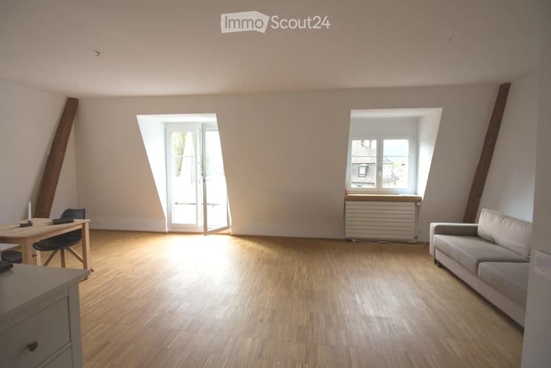 Bright, wooden-floored room with slanted ceilings, large windows, sofa, dining table, and chairs.