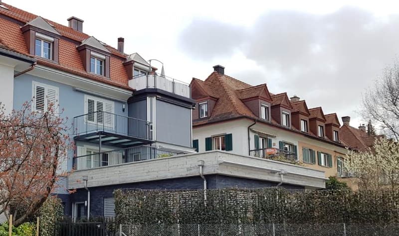 A multi-story residential building with a red-tiled roof, multiple windows, and balconies. The building features a mix of modern and traditional architectural elements, including a grey cylindrical tower and a white facade with green shutters.