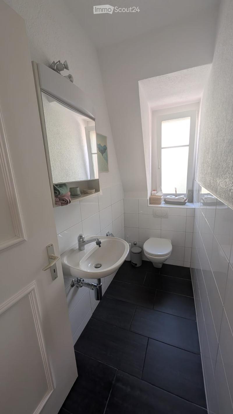 A small, modern bathroom with a white sink, chrome faucet, toilet, and a mirror with a shelf holding towels and a cup. The room has dark tiled flooring and white tiled walls, with a window providing natural light.