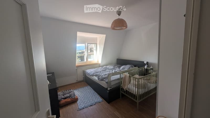 Cozy attic bedroom with wooden flooring, large window, bed, baby crib, rug, dog bed, and nightstand.