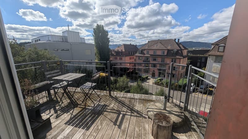 Balcony with wooden decking, metal railing, small table and chairs, and a tree stump as a seat.