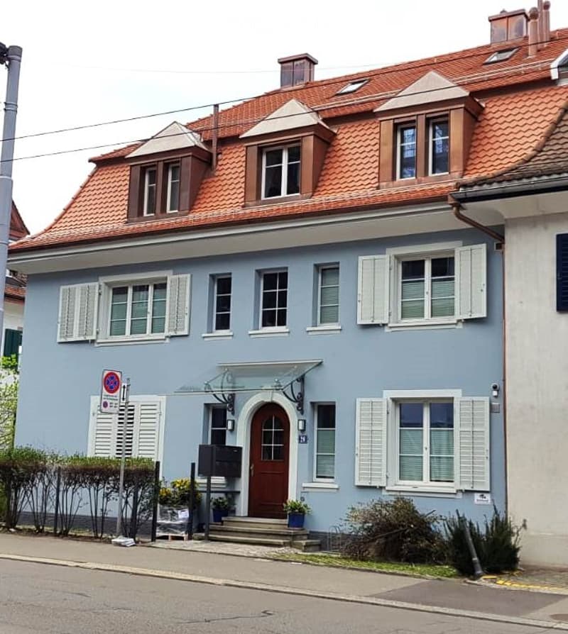 Two-story house with light blue exterior, red-tiled roof, white shutters, and a small porch with glass canopy above the entrance.