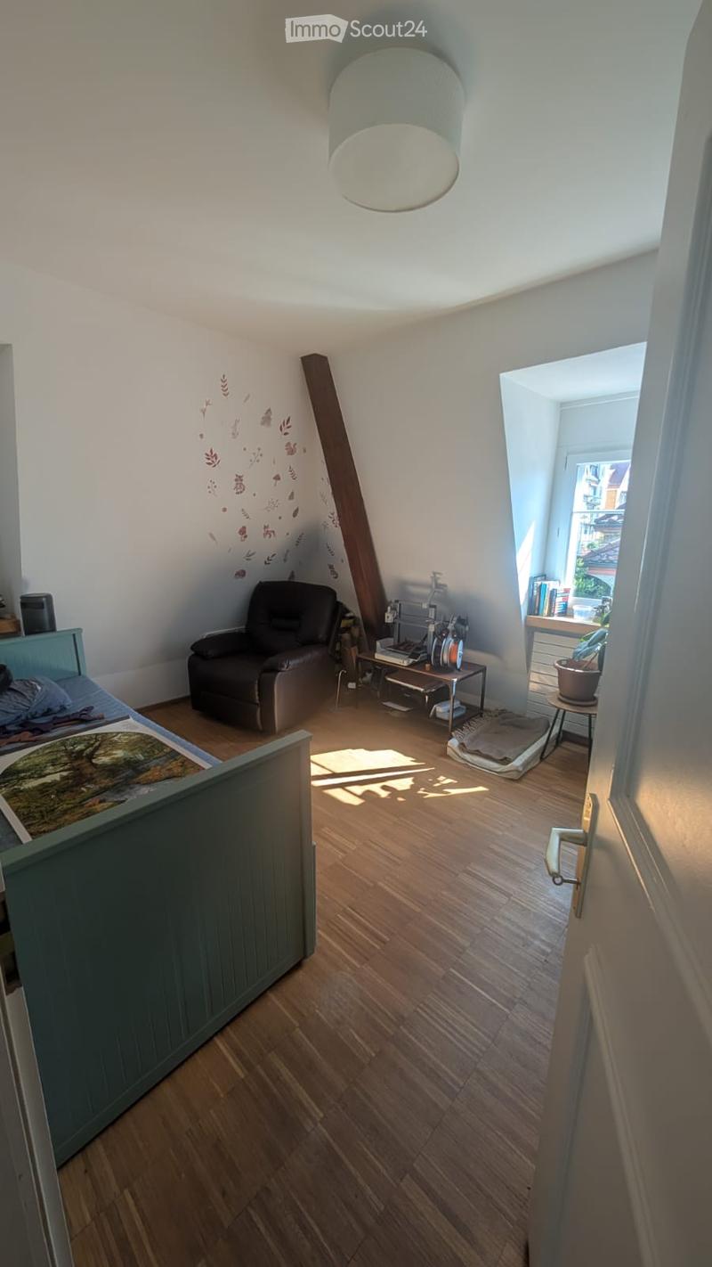 A cozy, well-lit room with wooden flooring, a large window, a comfortable sofa, a small table, a bookshelf, and a bed with a colorful blanket.