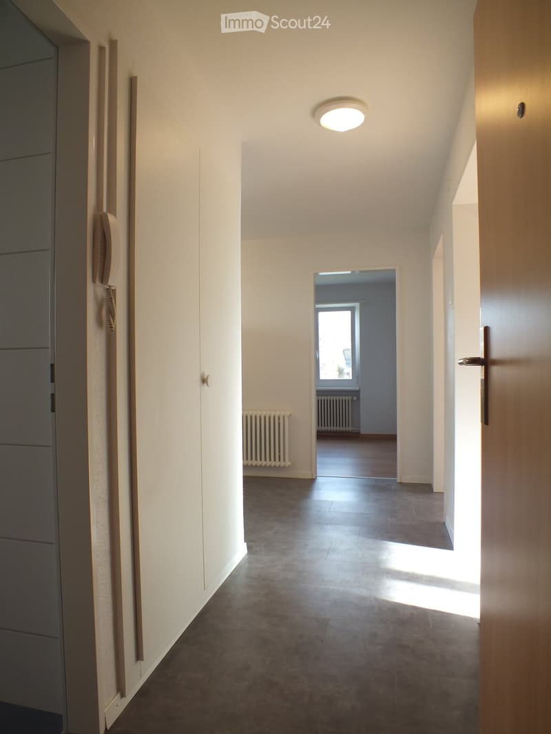 Empty hallway with white walls, grey tiled floor, and a ceiling light. A door leads to another room with a visible radiator and window.