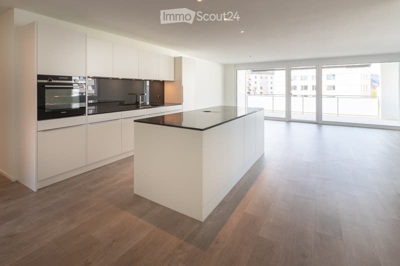 Modern kitchen with white cabinets, a central island, and a built-in oven. The space features wooden flooring and large windows offering a view of the city.