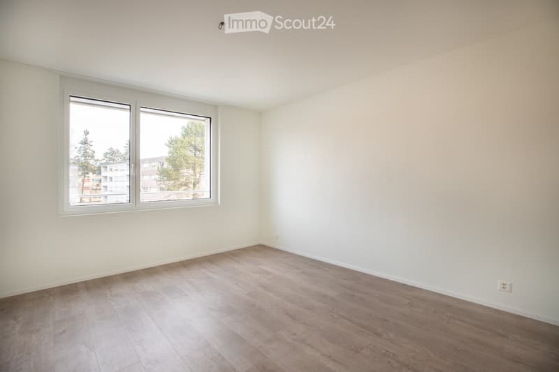 Empty room with light wooden flooring and white walls, featuring large windows with white frames and a view of trees and buildings outside.
