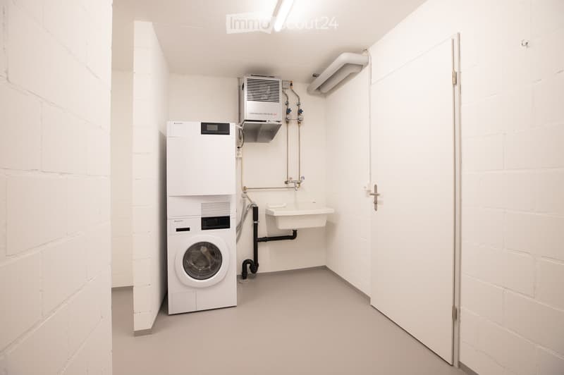 A small, clean utility room with white brick walls and a white floor. It contains a washing machine, a water heater, and a small sink.