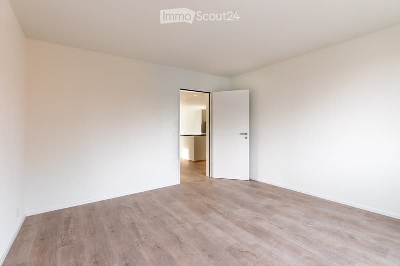 Empty room with light-colored walls and wooden flooring, featuring an open doorway leading to another room.