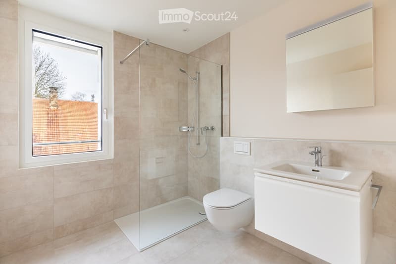 Modern bathroom with a glass-enclosed shower, toilet, sink, and large mirror. The room features beige tiles and a large window offering a view of the garden.