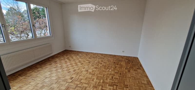 Empty room with wooden herringbone flooring, white walls, and large windows with a view of trees.
