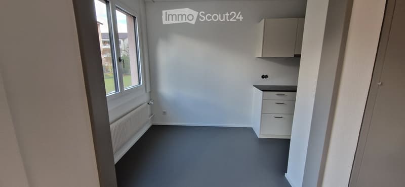 Empty room with white walls, grey flooring, a window with a view, a radiator, and a small cabinet with drawers.