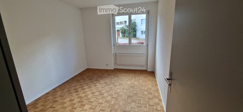 Empty room with wooden parquet flooring, white walls, a window with white frame, and a radiator below the window.