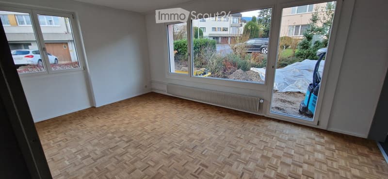 Empty room with wooden parquet flooring, large windows, radiator, and a sliding glass door leading to an outdoor area.
