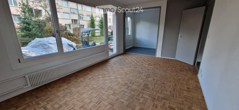 Empty room with wooden herringbone flooring, large windows, and a radiator. No furniture or equipment present.