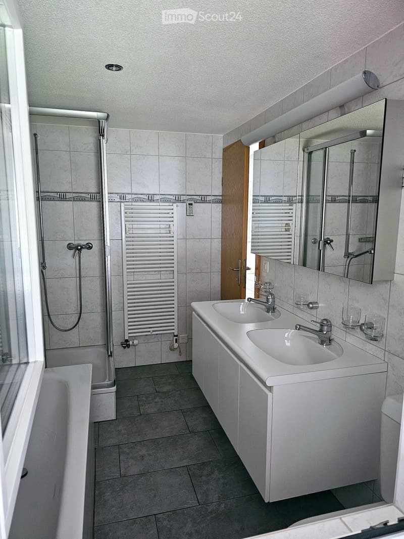 Modern bathroom with double sink, large mirror, bathtub, and shower area. Features tiled walls and floor, radiator, and glass shower enclosure.