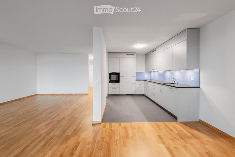 Empty room with light wooden flooring, white walls, and a small kitchen area featuring white cabinets, a sink, and an oven. No furniture or personal items are present.