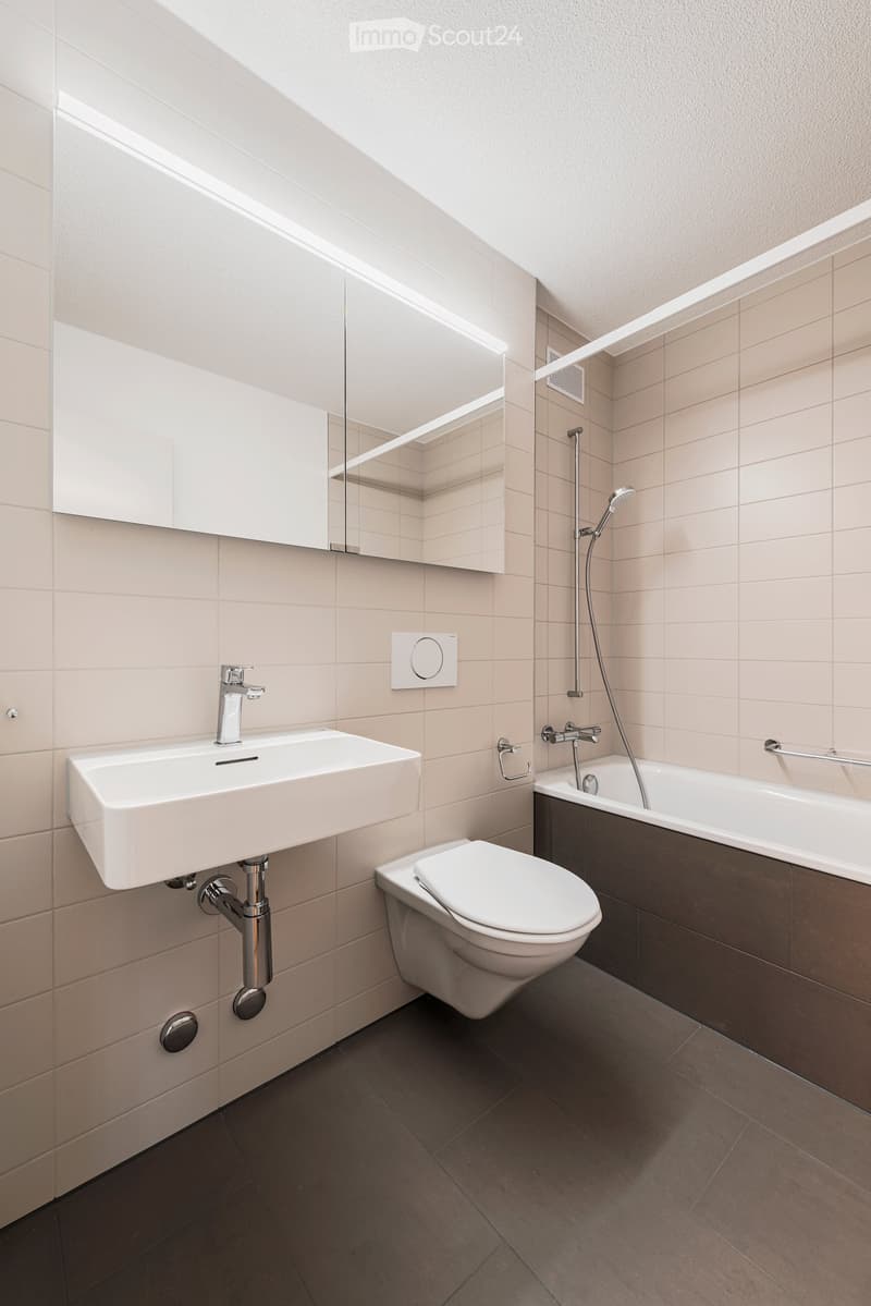 Modern bathroom with a white sink, chrome faucet, toilet, bathtub with showerhead, and a mirrored cabinet above the sink. Light beige tiles on walls and dark tiles on the floor.