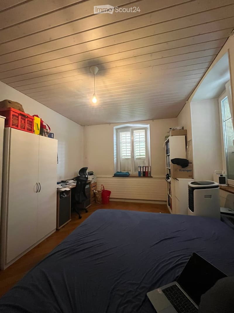 A furnished bedroom with a bed, desk, chair, laptop, and various storage items including a cabinet, refrigerator, and bookshelf. The room has wooden flooring and white walls with a slanted ceiling.
