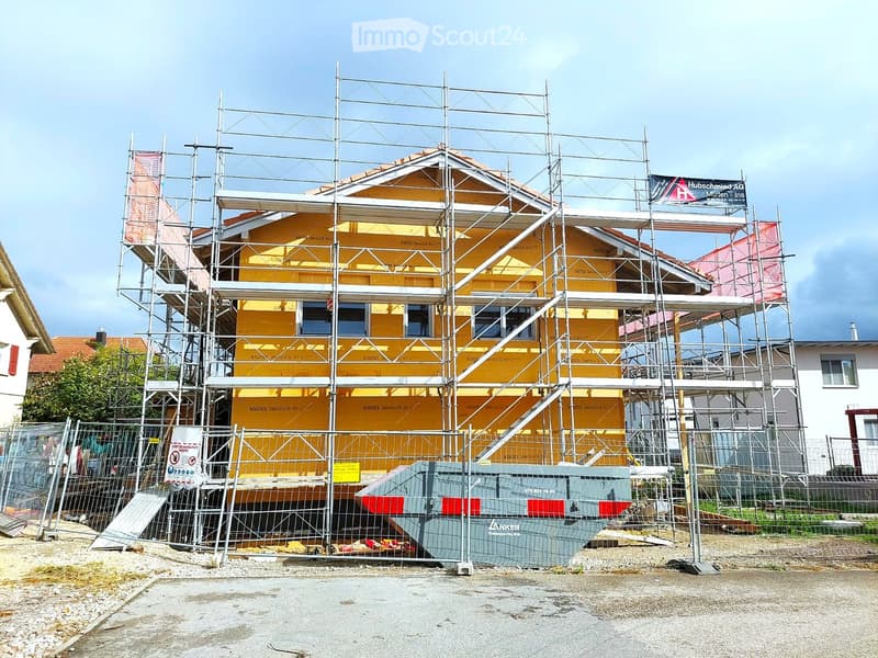 A two-story house under construction, covered in scaffolding with yellow exterior walls visible. The building is enclosed by a metal fence with signage and construction materials nearby.