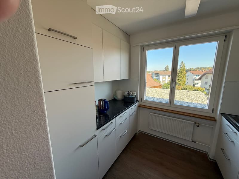 Modern kitchen with white cabinets, dark countertop, coffee maker, and a large window overlooking a residential area.