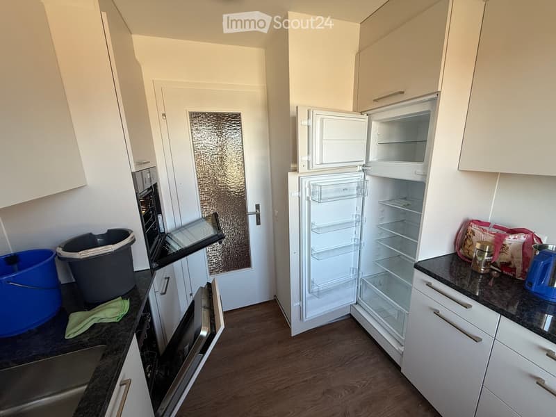 Modern kitchen with white cabinets, open refrigerator, oven, countertop, and various items including a cup and trash bins.