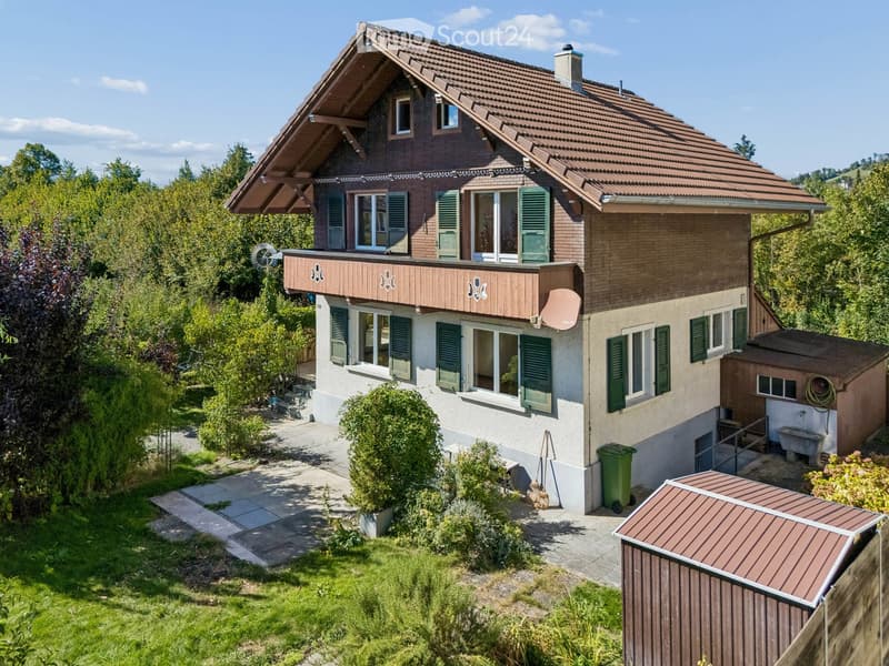 Two-story house with a tiled roof, green shutters, and a wooden balcony. The house is surrounded by a well-maintained garden with trees and shrubs.