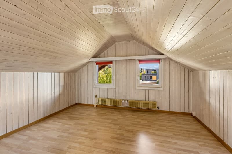 Empty room with wooden walls, wooden floor, and two windows with blinds. A radiator is visible under the windows.