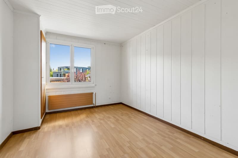 Empty room with wooden flooring, white paneled walls, and a large window offering a view of the outside. A radiator is visible below the window.