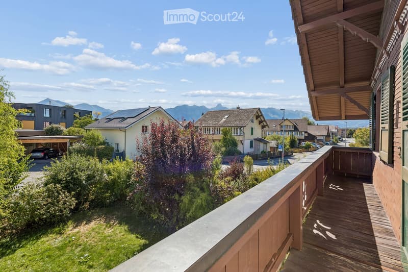 Balcony overlooking a residential area with houses, greenery, and distant mountains under a clear blue sky.