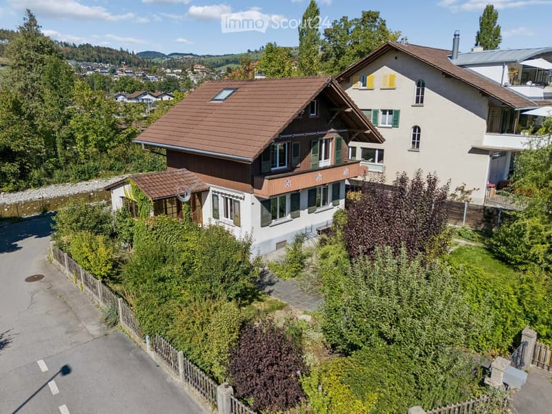 A two-story house with a red-tiled roof, green shutters, and a well-maintained garden. The house features multiple windows and a small balcony on the upper floor.