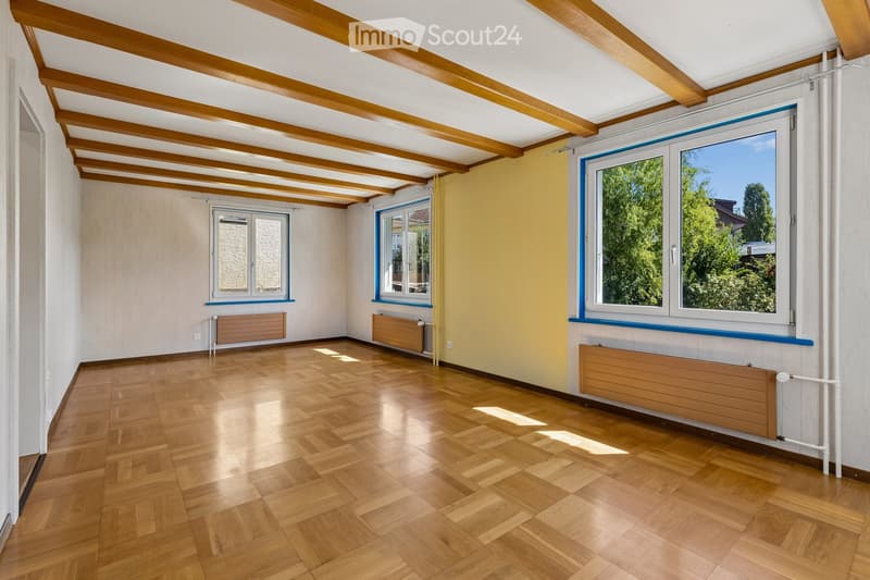 Empty room with wooden flooring, exposed wooden beams on the ceiling, and large windows with blue frames. The walls are painted in neutral tones, and there are radiators along the walls.