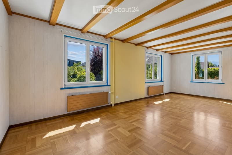 Empty room with wooden flooring, exposed wooden beams on the ceiling, large windows with blue frames, and radiators along the walls.