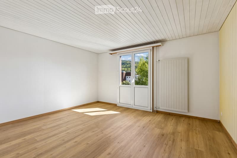 Empty room with wooden flooring, white walls, and a large window with a view of greenery outside.