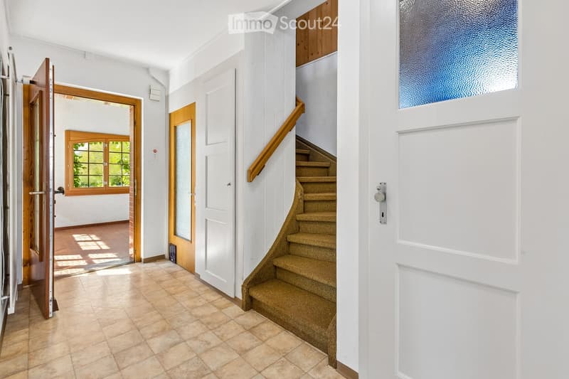 Bright hallway with tiled floor, white walls, wooden doors, and a carpeted staircase leading upwards.