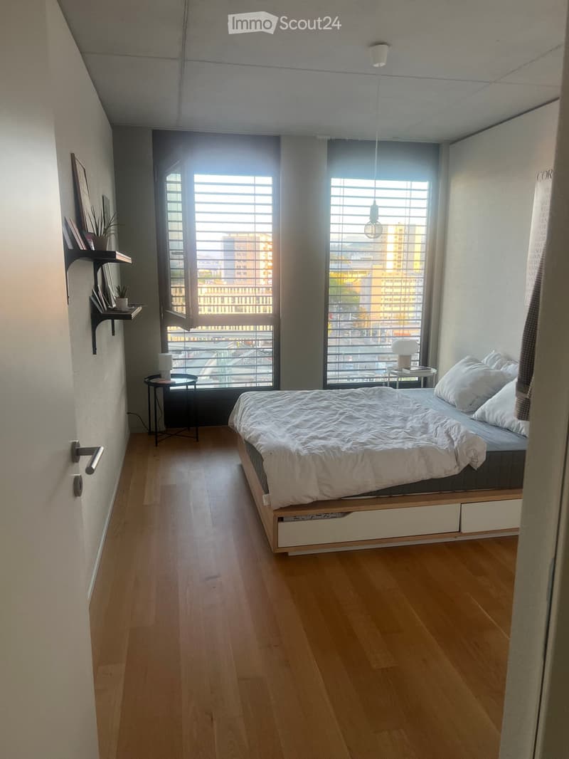 Modern, minimalist bedroom with wooden flooring, large windows with blinds, a bed with white bedding, a nightstand, a small table, a shelf with books and a plant, and a hanging light fixture.