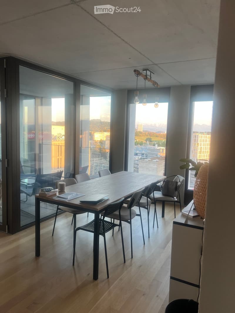 Bright, modern room with large windows offering a city view, featuring a wooden table surrounded by chairs, a vase with plants, and a few books.
