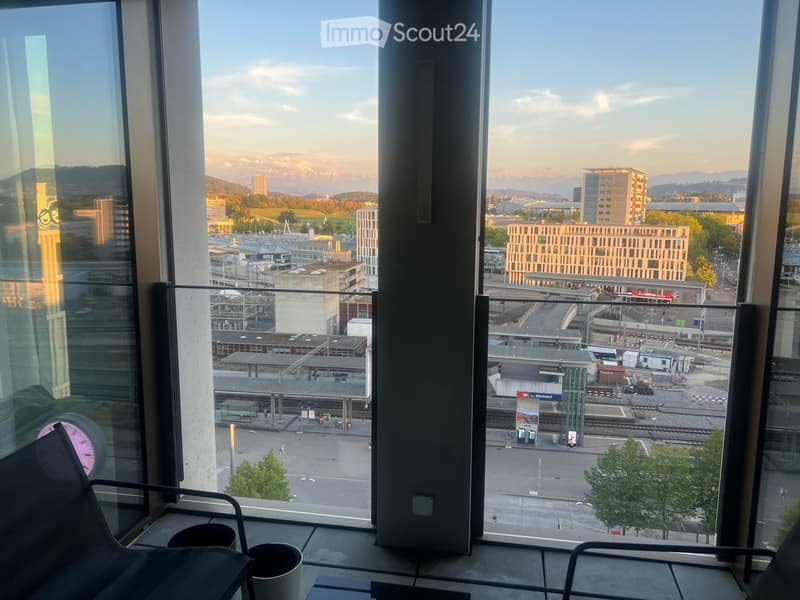 A modern room with large windows offering a panoramic view of a cityscape, including buildings, a train station, and distant mountains. The room features contemporary furniture, including a black chair and a small table.