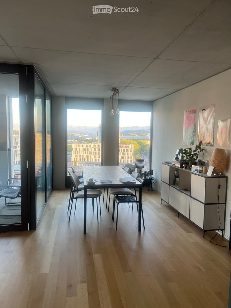 Bright, modern room with wooden flooring, large windows, a dining table with chairs, a cabinet, and a lamp.