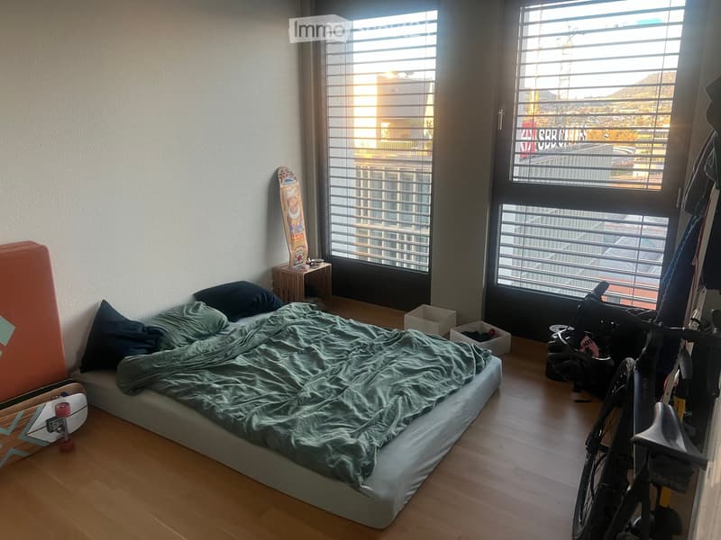 A small, modern bedroom with a single bed, nightstand, skateboard, bicycle, and storage boxes. Large windows offer a view of the city.
