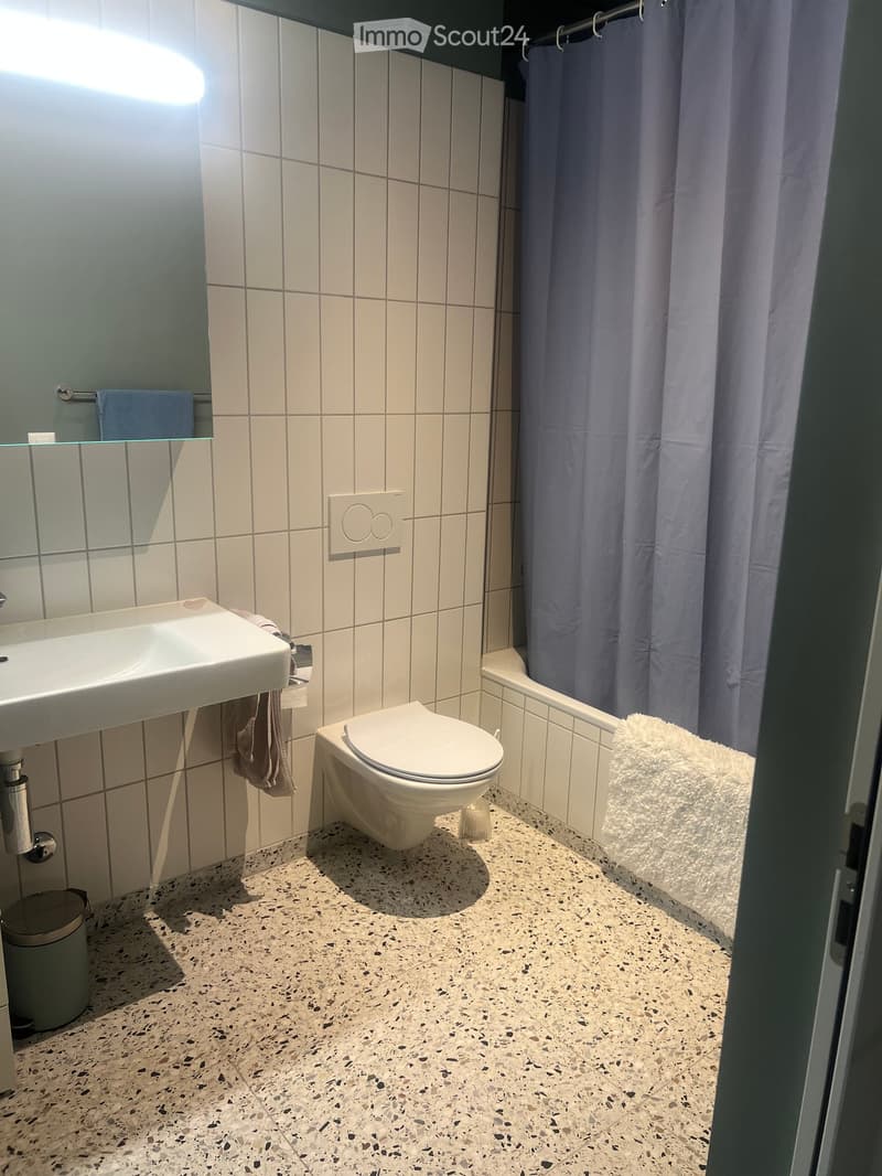 Modern bathroom with white tiles, a sink, toilet, shower curtain, and a towel on the edge of the bathtub.