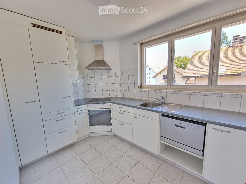 Modern kitchen with white cabinets, stainless steel appliances, and large windows overlooking a residential area.