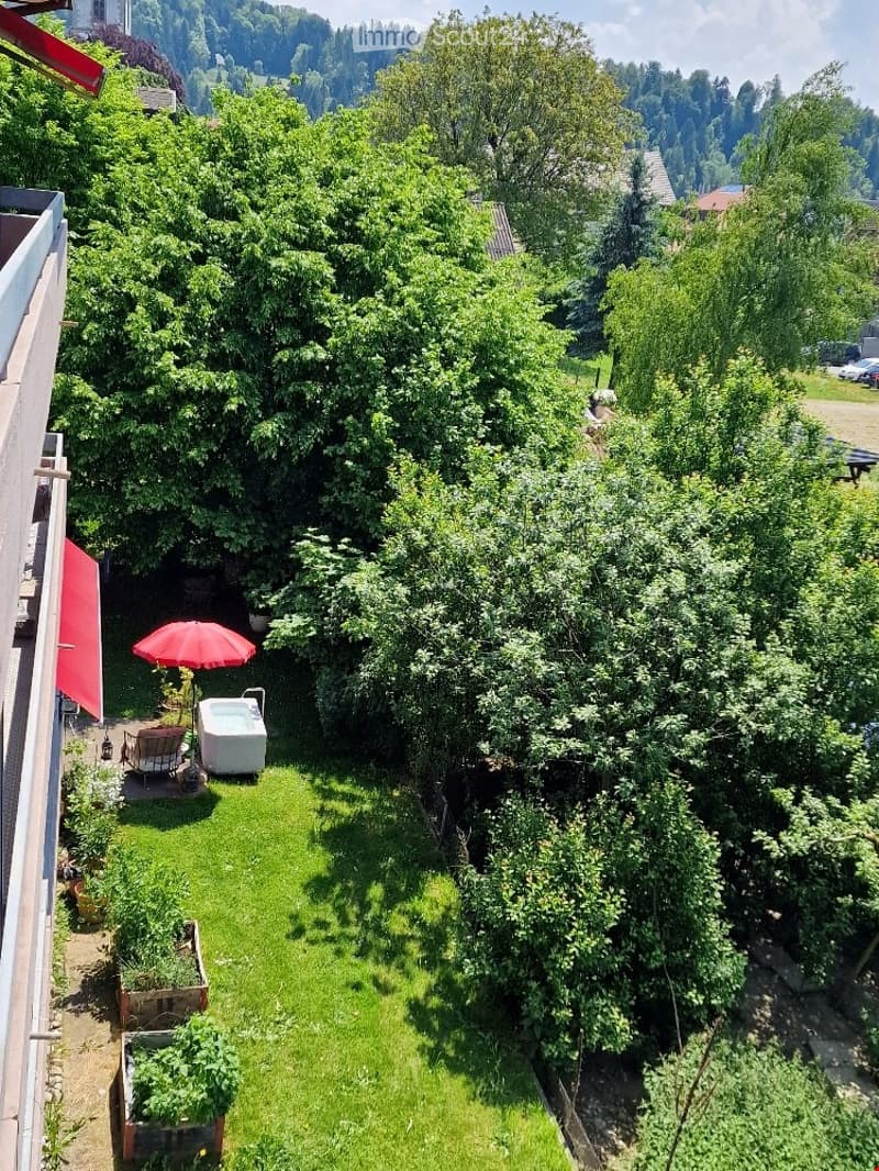 A lush garden with a small patio area, featuring a red umbrella, a small table, and a hot tub, surrounded by trees and greenery with a scenic view of the landscape.