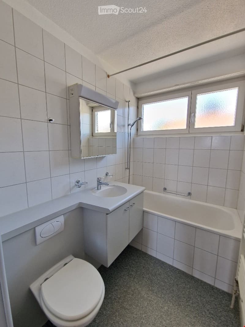 Modern bathroom with white tiles, a sink with a mirror cabinet, a bathtub, and a toilet. Natural light from a window illuminates the space.