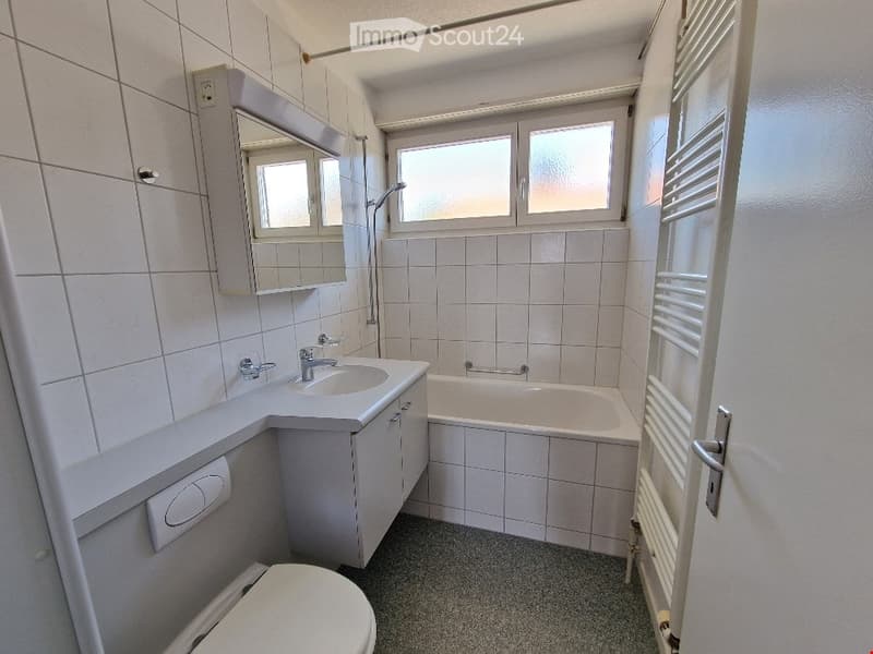 Modern bathroom with white tiles, a sink with a mirror cabinet, a bathtub, a toilet, and a radiator next to the window.